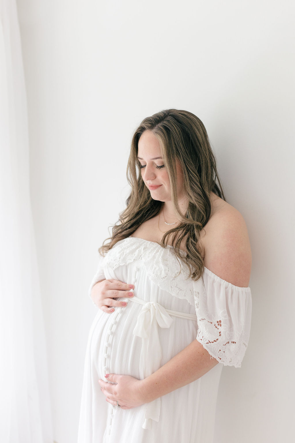 A mom to be leans on a wall while smiling down at her bump in a white maternity gown