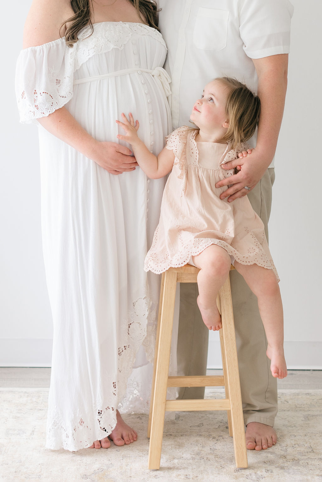 A toddler girl in a peach dress sits on a wooden stool looking up to mom and dad behind her