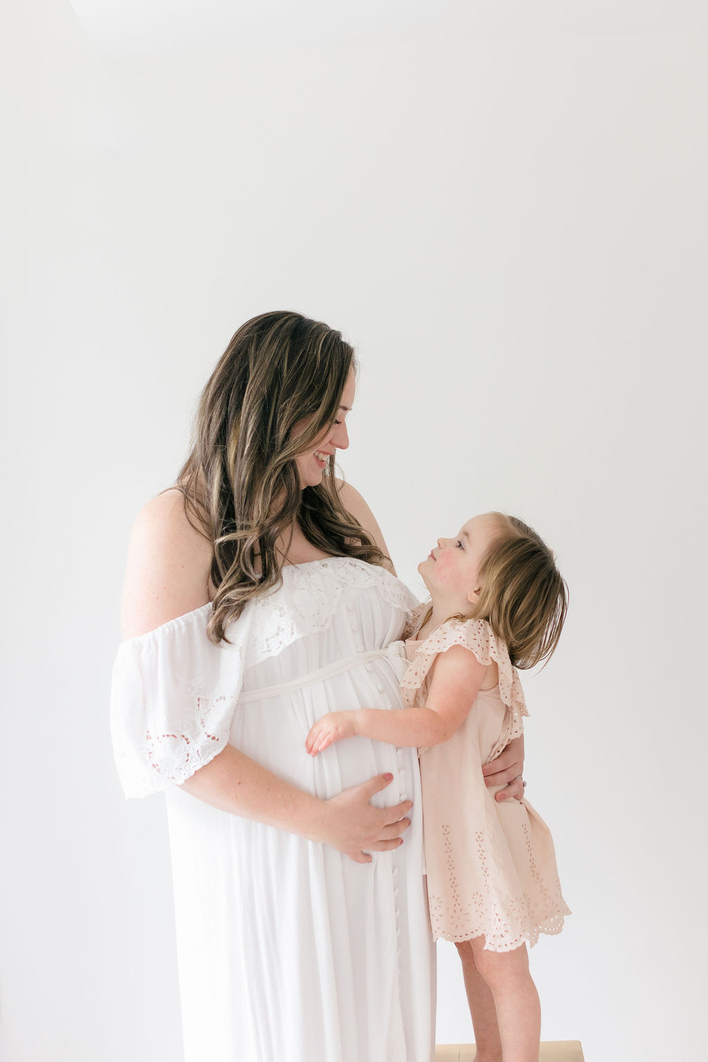 A happy pregnant mother hugs her toddler daughter standing on a stool in a studio