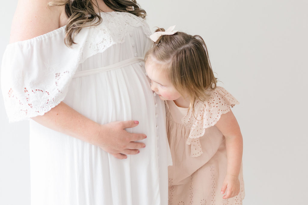 A toddler girl kisses mom's pregnant bump while in a studio in a white maternity dress from one of the shops for maternity clothes philadelphia