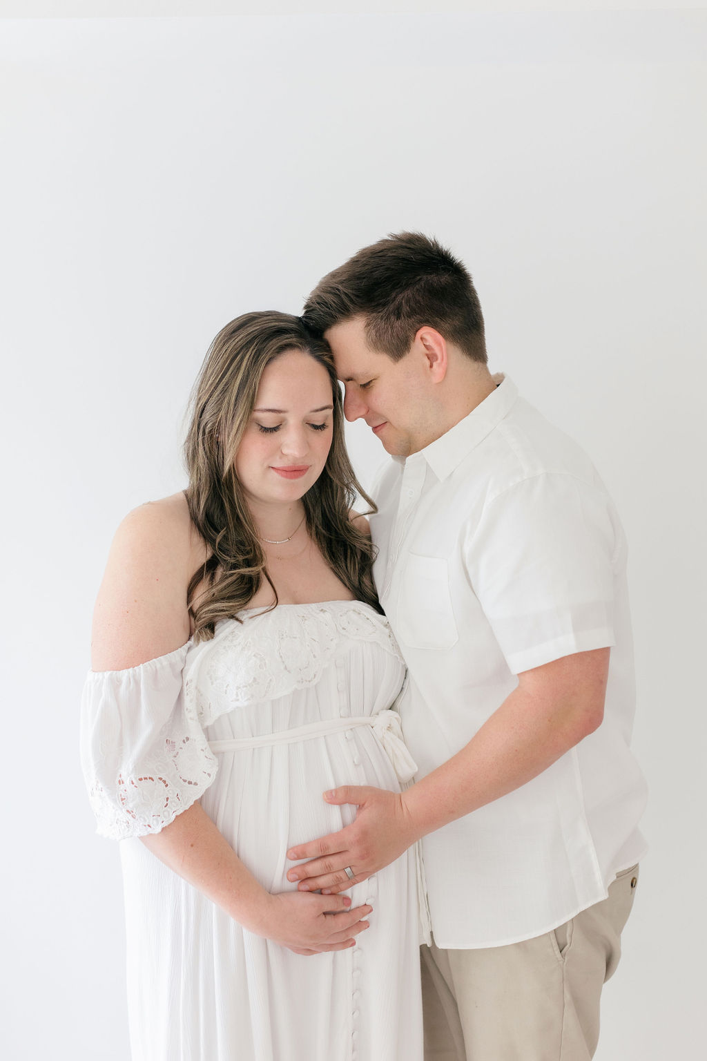 Happy expecting parents stand in a studio snuggling while holding the bump after shopping for maternity clothes philadelphia