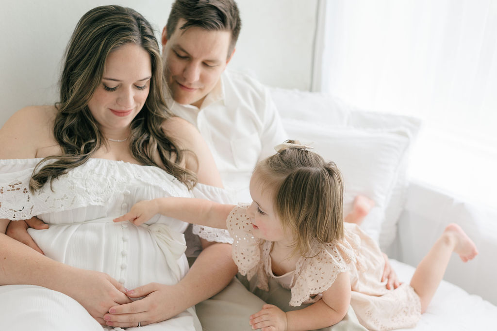 A toddler girl in a cream dress points to mom's pregnant bump while playing on a bed after finding maternity clothes philadelphia