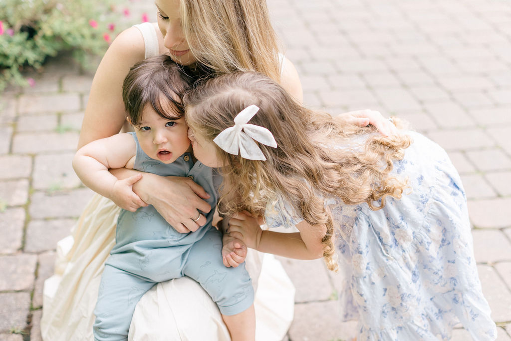 A toddler girl in a blue dress kisses the cheek of her baby brother in mom's lap in a garden patio