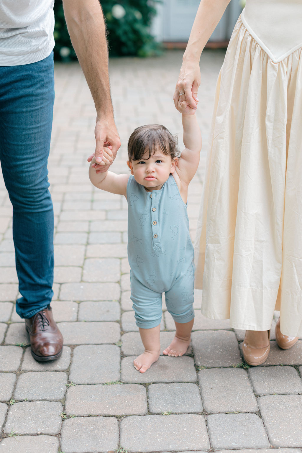 A grumpy toddler in a blue onesie walks with mom and dad's help on a patio before visiting pumpkin patches philadelphia