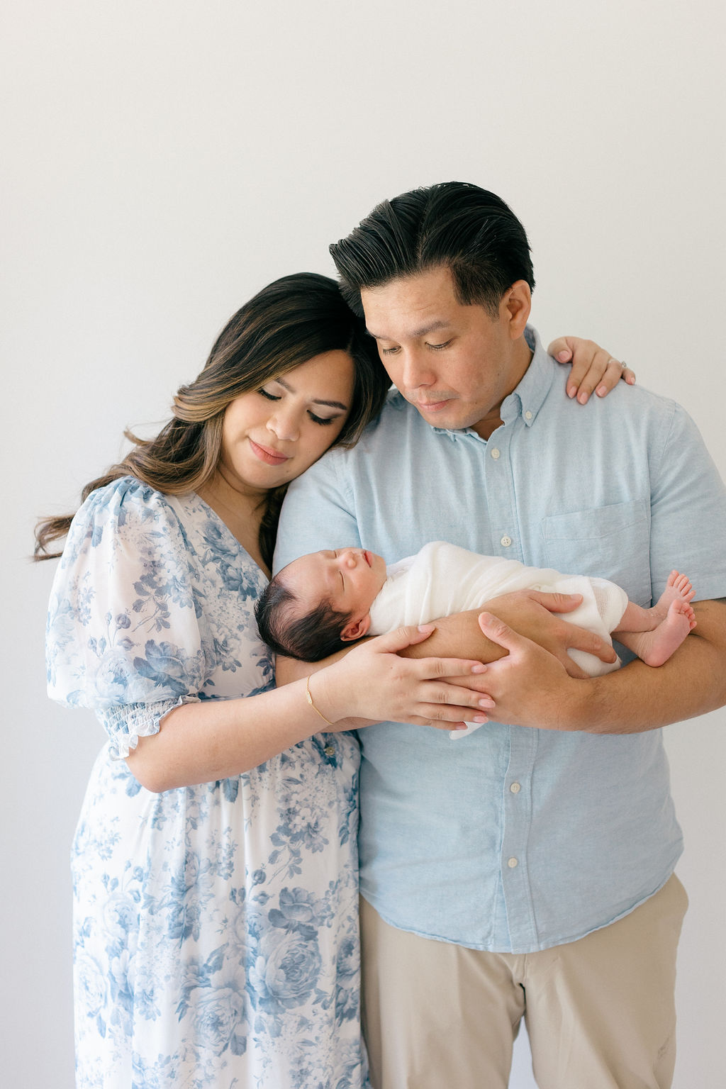 New parents cuddle while standing in a studio holding their sleeping newborn baby