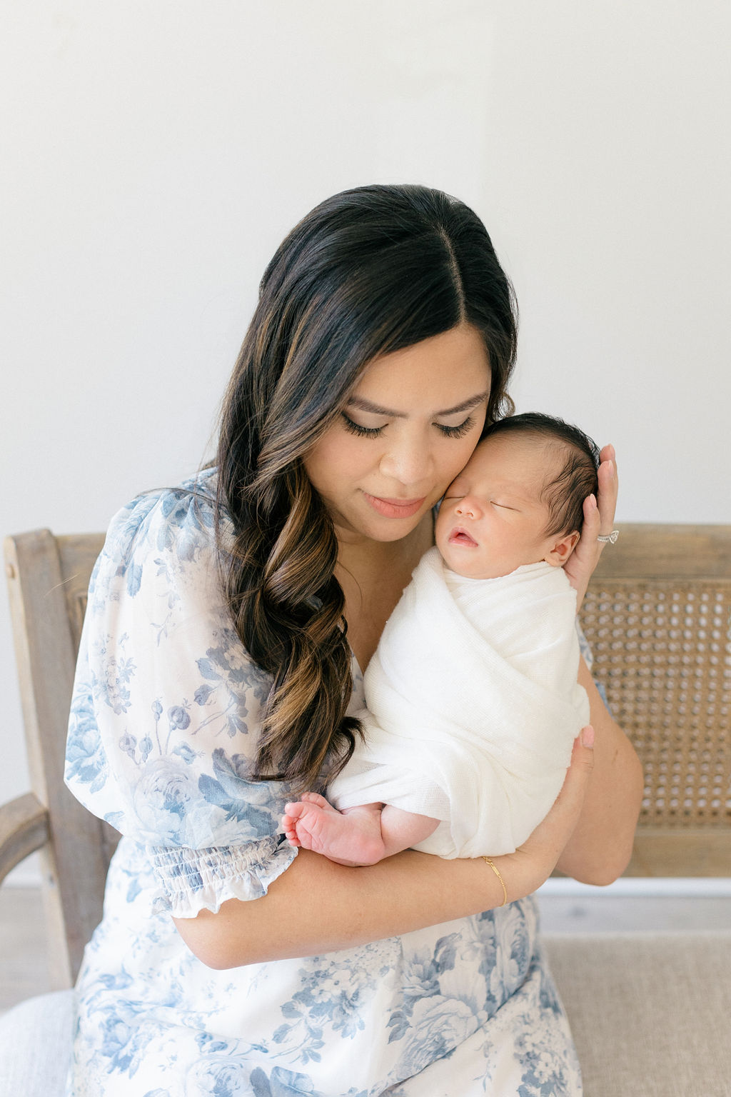 A mother cradles her sleeping newborn baby against her cheek while sitting on a bench in a studio