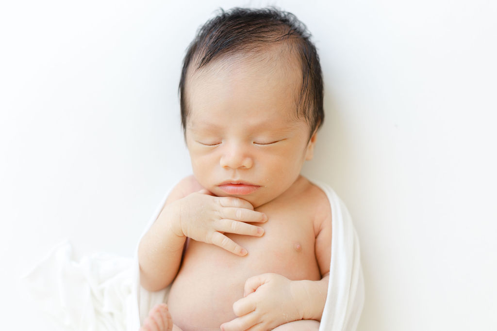 A newborn baby sleeps with a hand under its chin in a studio on a white bed