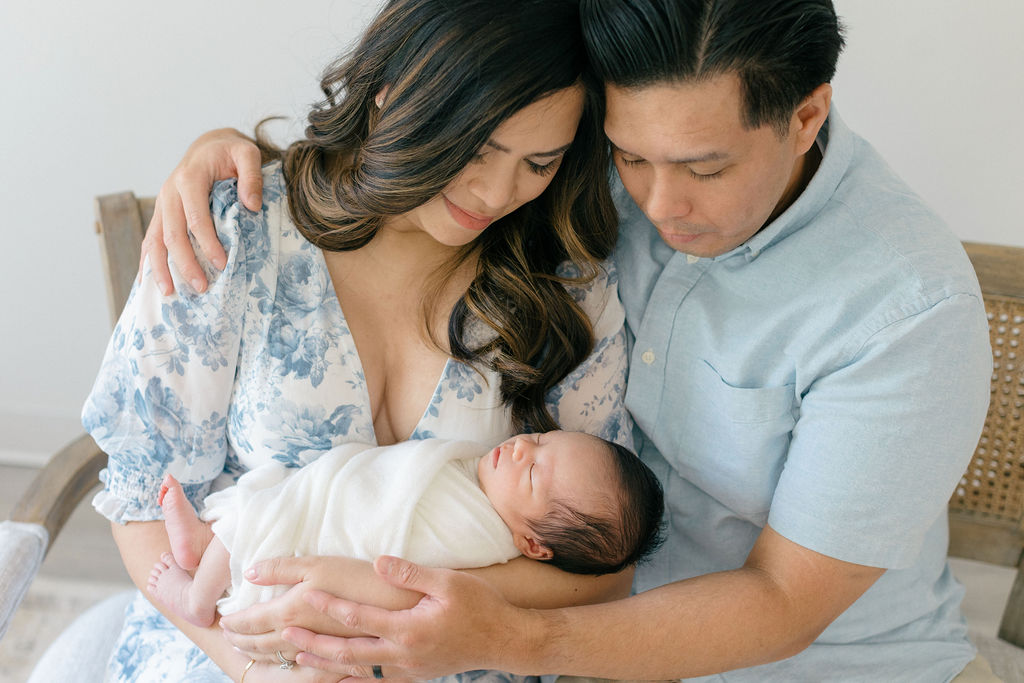 New parents sit on a bench in a studio cradling their sleeping newborn baby in their laps before visiting toy stores main line pa