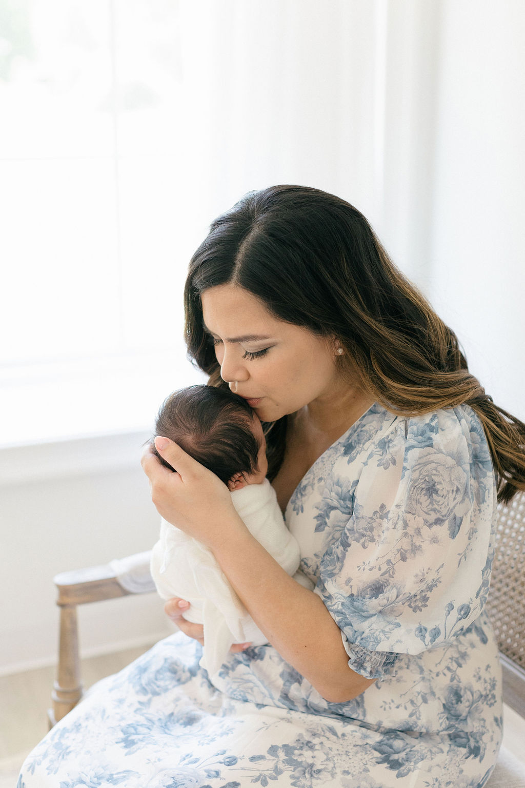 A happy mother kisses the head of her newborn baby while sitting on a bench in a studio window before visiting toy stores main line pa