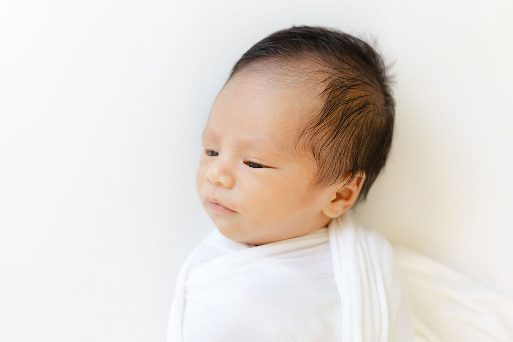 A newborn baby laying on a white bed in a white swaddle with eyes open before visiting toy stores main line pa