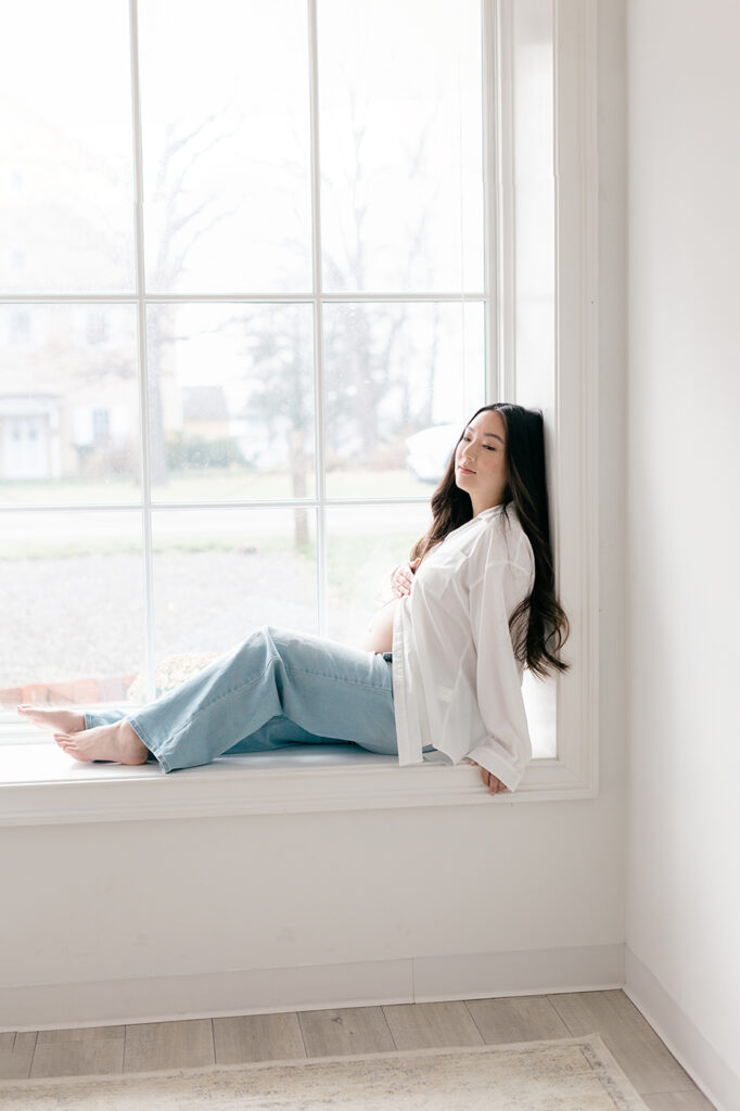 mom-to-be sitting on window jeans white open top