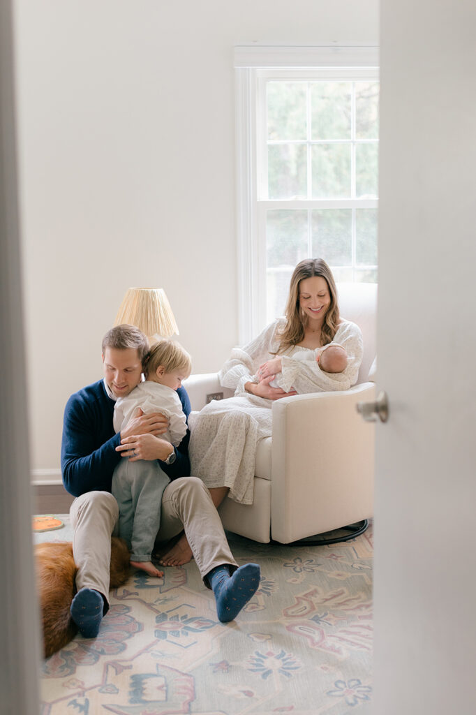 family of four in nursery, mom rocking newborn and dad hugging toddler boy