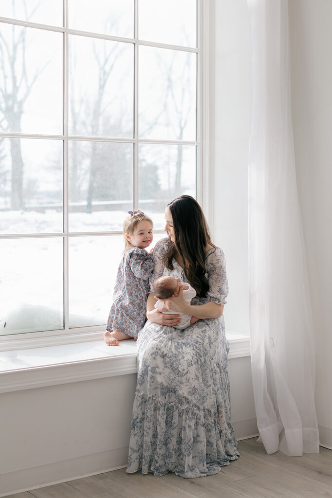 mom baby and toddler sitting on a window seat smiling philadelphia studio newborn session